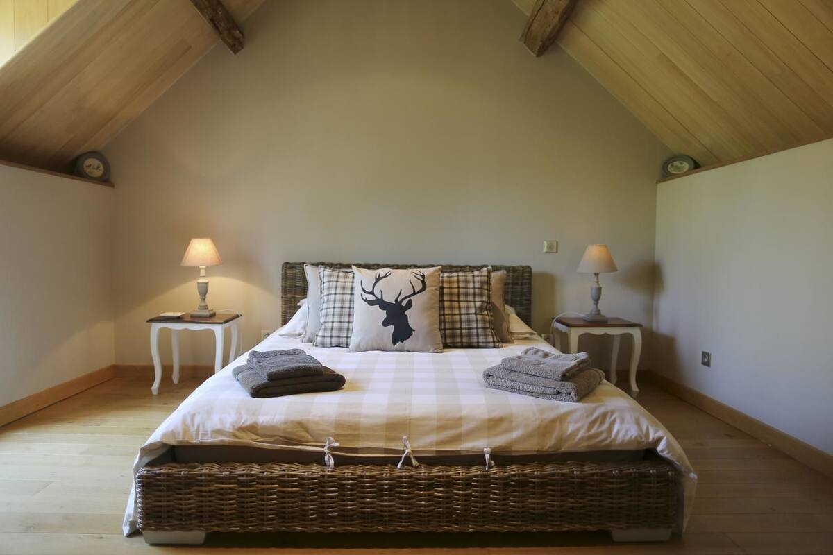 A bedroom with a high ceiling, a large bed with a checkered bedspread, two nightstands with lamps, and a decorative pillow with a deer silhouette.