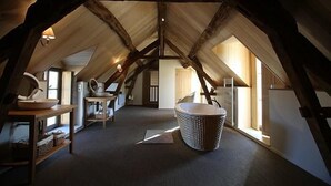Une chambre située sous les combles, avec des poutres apparentes, une suspension décorative et une grande fenêtre laissant entrer la lumière naturelle.