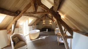 Une pièce mansardée avec des poutres apparentes, un fauteuil rembourré, un panier en osier et une petite table.