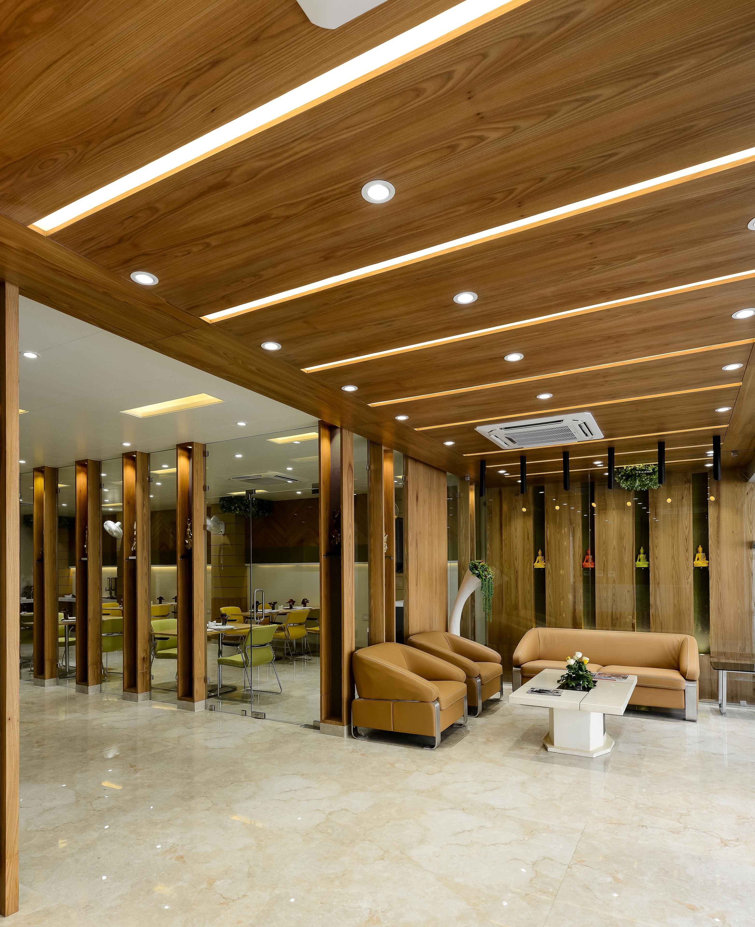 A modern lobby with wooden paneling, recessed lighting, a seating area with a sofa and armchairs, and a glass partition separating the seating from the dining area.