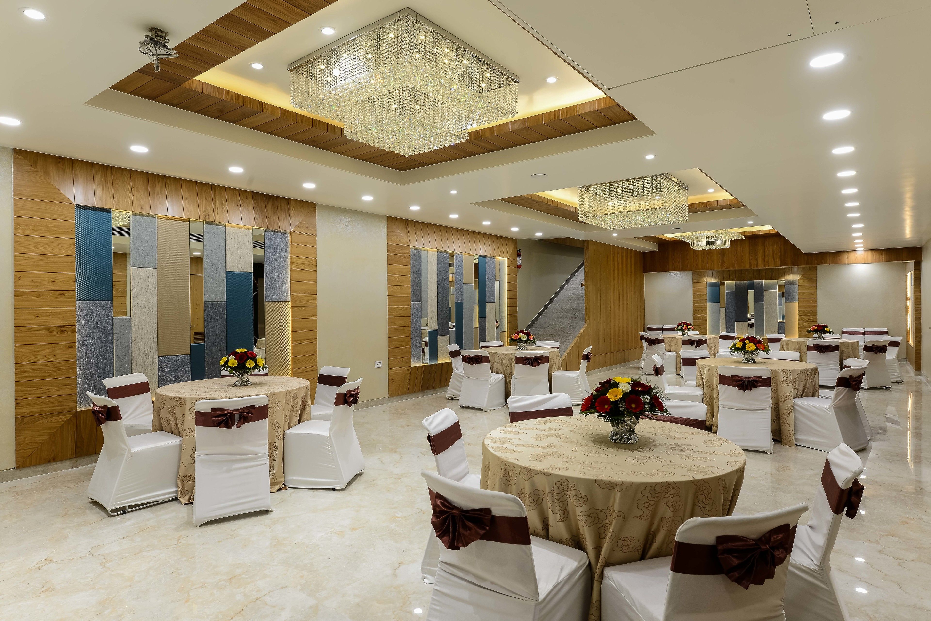 A banquet hall with round tables, white chairs with brown sashes, and chandeliers.