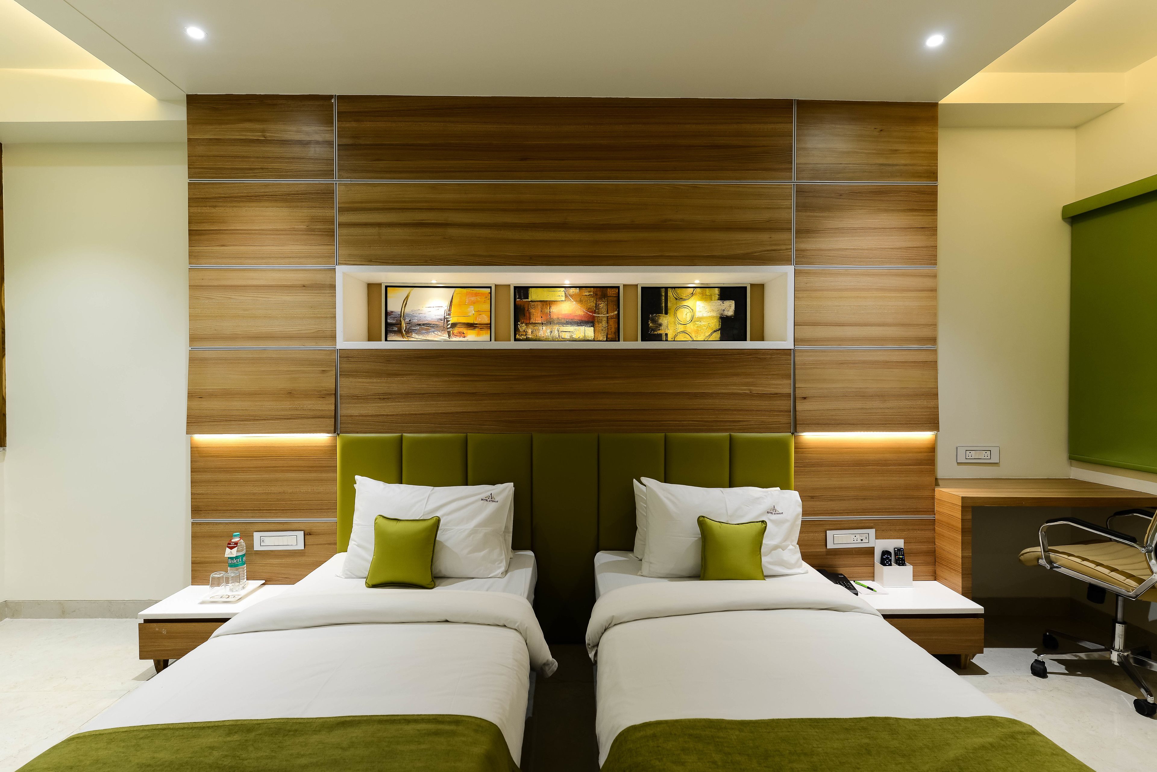 A hotel room with two beds, a wooden headboard, a green accent wall, and a small shelf with artwork.