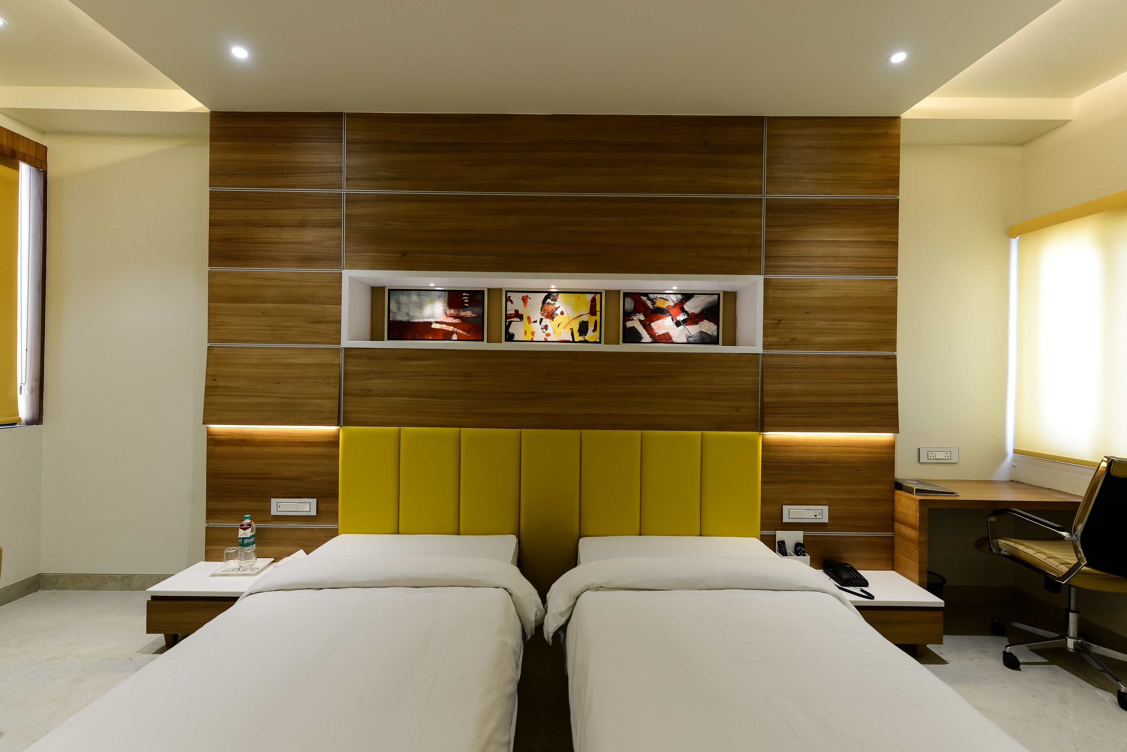 A hotel room with two beds, a wooden headboard, a desk, and a chair. There are three paintings on the wall above the beds.