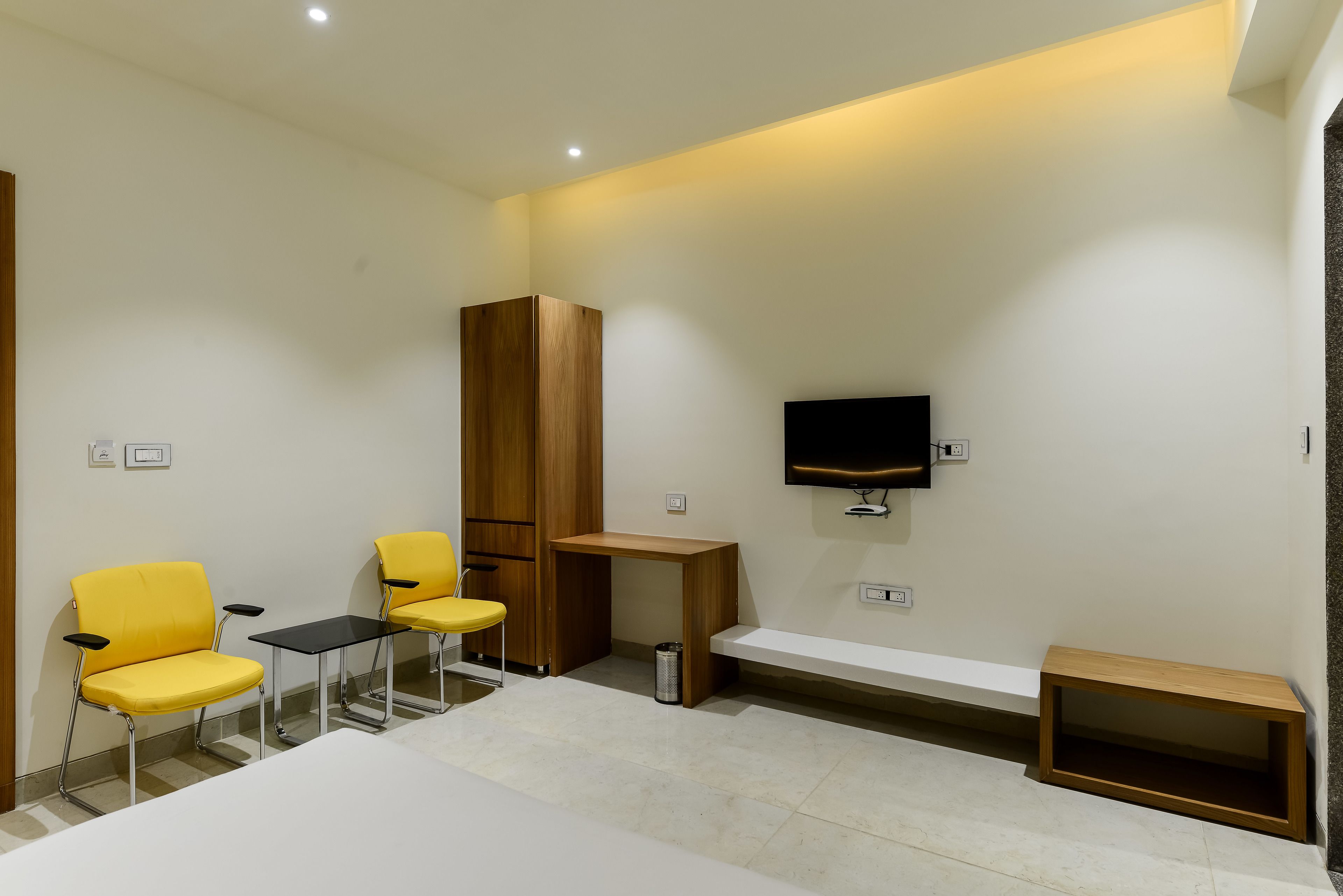 A modern hotel room with a bed, two yellow chairs, a small table, a wooden cabinet, a TV mounted on the wall, and a bench.