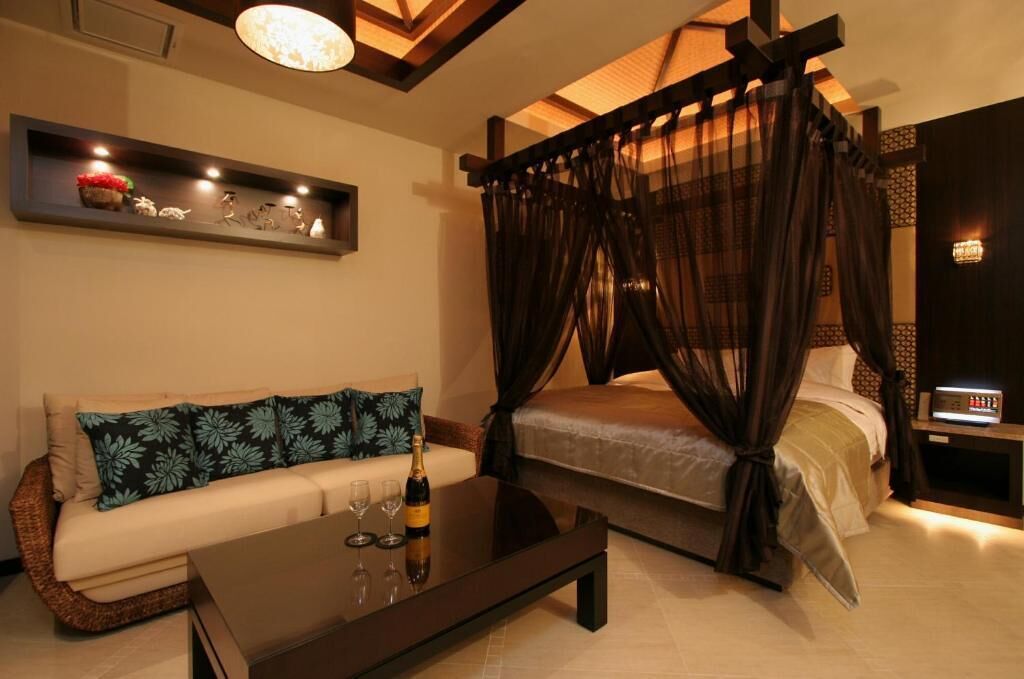 A bedroom with a four-poster bed, a sofa, a bedside table with a lamp, and a wall-mounted shelf with decorative items.
