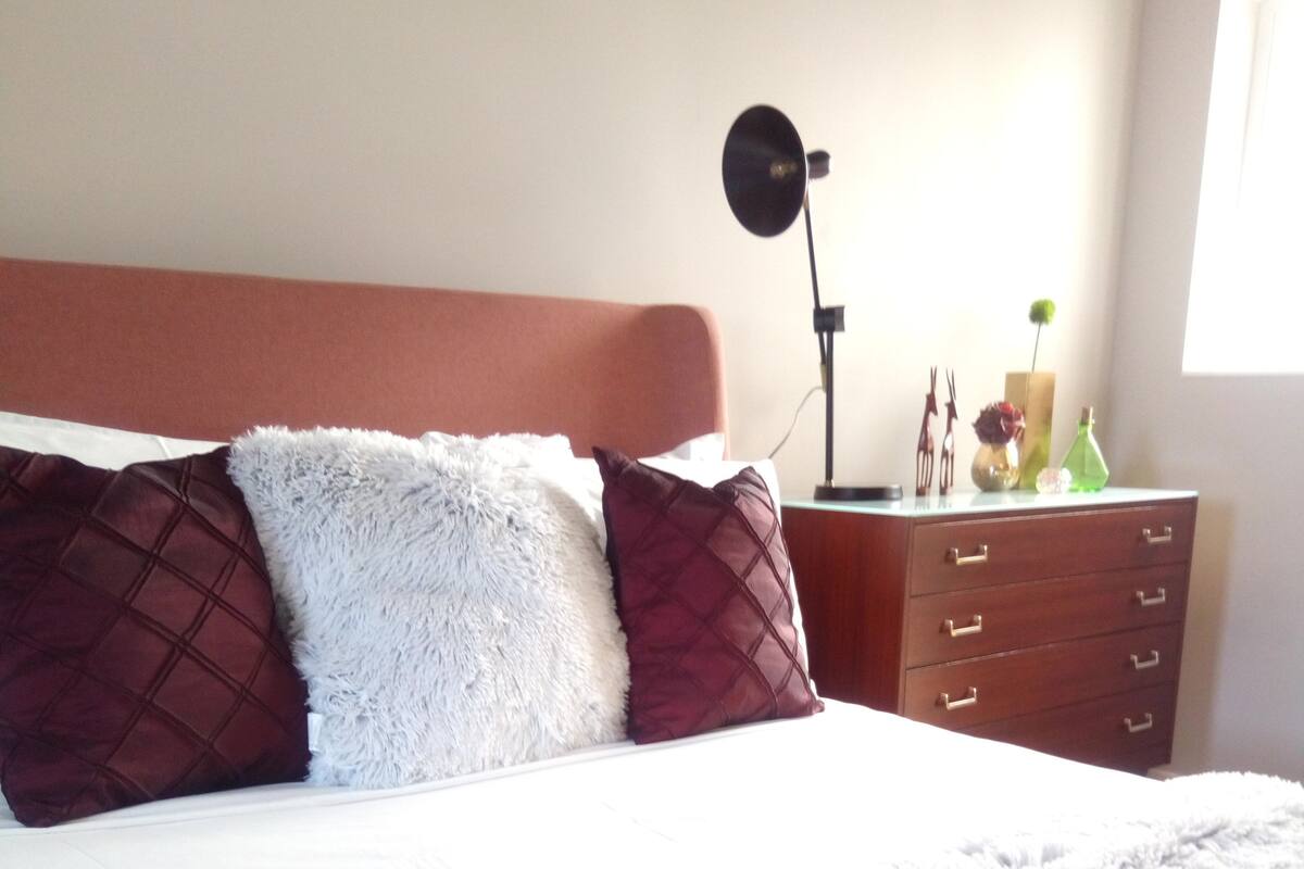 A neatly made bed with a fluffy white blanket and two pillows, a bedside table with a lamp, and a dresser with drawers.