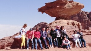 A group of people sitting on a sandy area with a large rock formation in the background. - Mars Private camp (Wadi Rum)