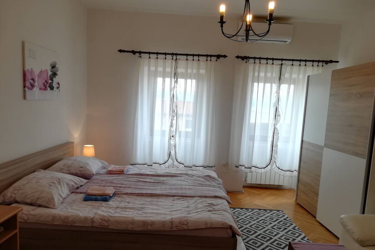 A bedroom with a bed, a nightstand, a lamp, curtains, a chandelier, and a wall-mounted air conditioner.