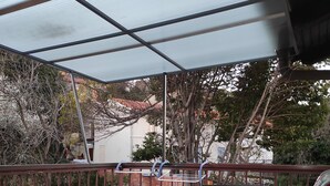 An outdoor patio with metal chairs and a table, a covered area, and a view of a residential area with trees and buildings. - Apartments Patricija (Opatija)