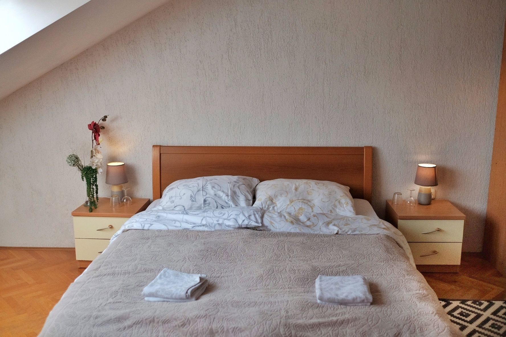 Ein ordentlich bezogenes Bett mit einem hölzernen Kopfteil, zwei Nachttische mit Lampen und eine Blumenkomposition auf einem Tisch.