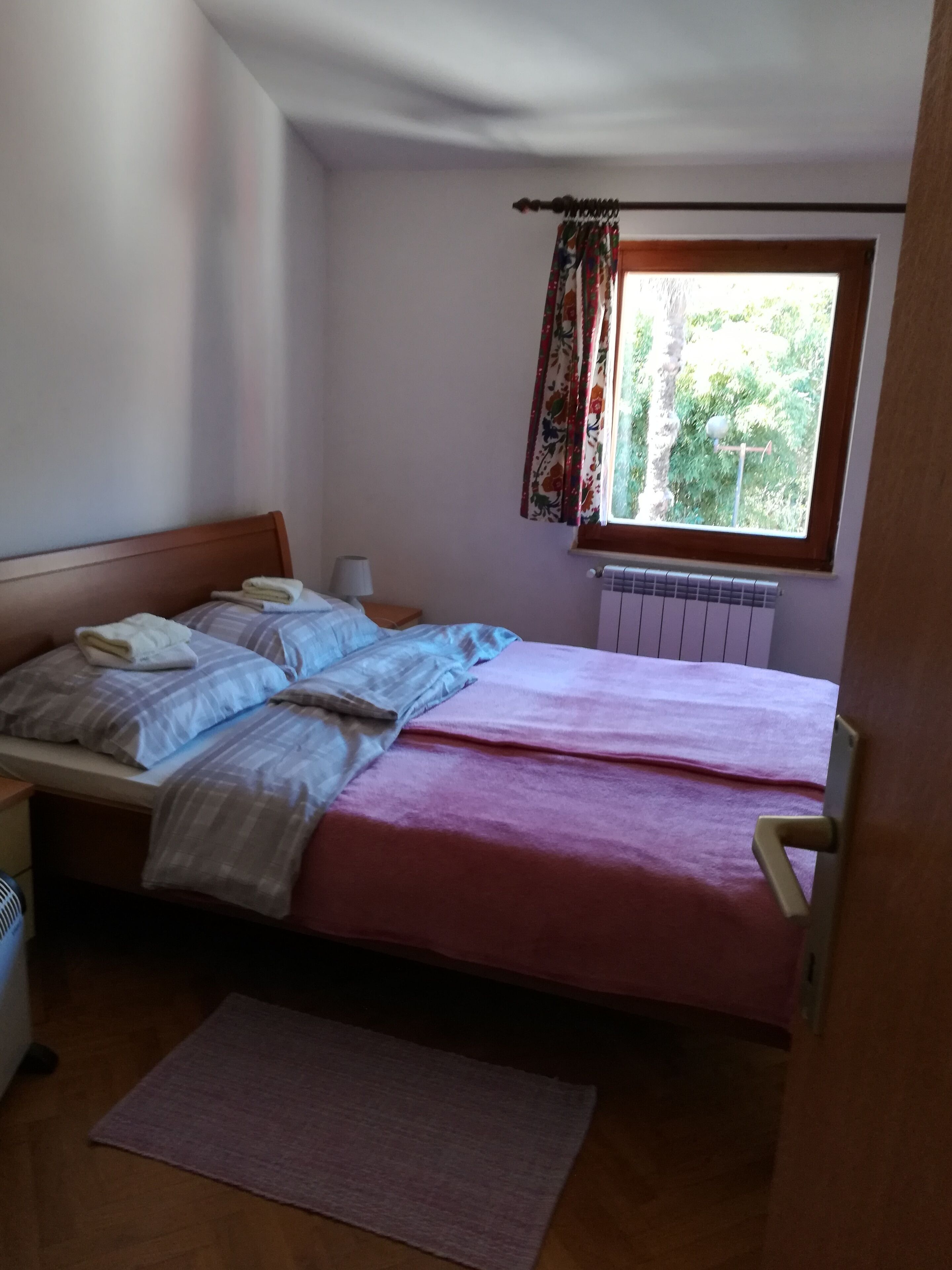 A bedroom with a double bed, a bedside table, a lamp, a radiator, and a window with curtains.