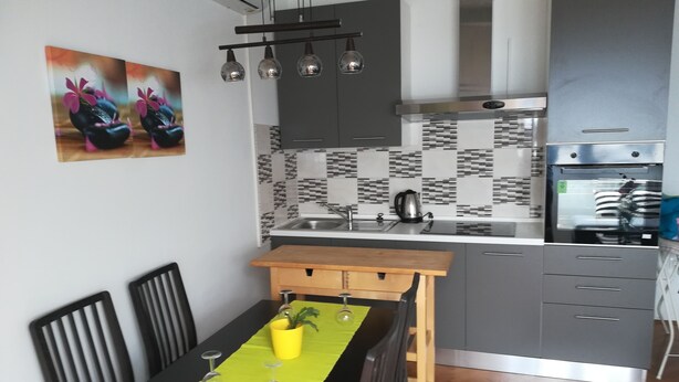 A modern kitchen with grey cabinets, a black countertop, and a tiled backsplash. There is a dining table with chairs and a yellow placemat. - Apartments Patricija (Opatija)