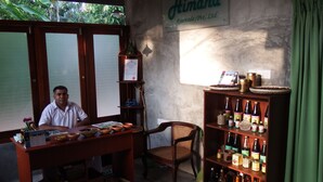 A man sitting at a wooden desk with a shelf of bottles, a framed certificate, and a sign reading "Himana Ayurveda (Pvt) Ltd" on the wall. - Himana Ayurveda (Seeduwa - Katunayake)