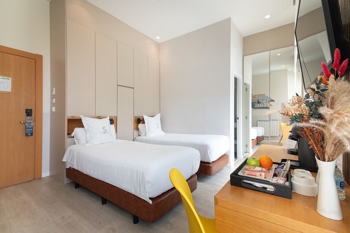A hotel room with two beds, a wooden desk, a mirror, and a vase with dried plants.