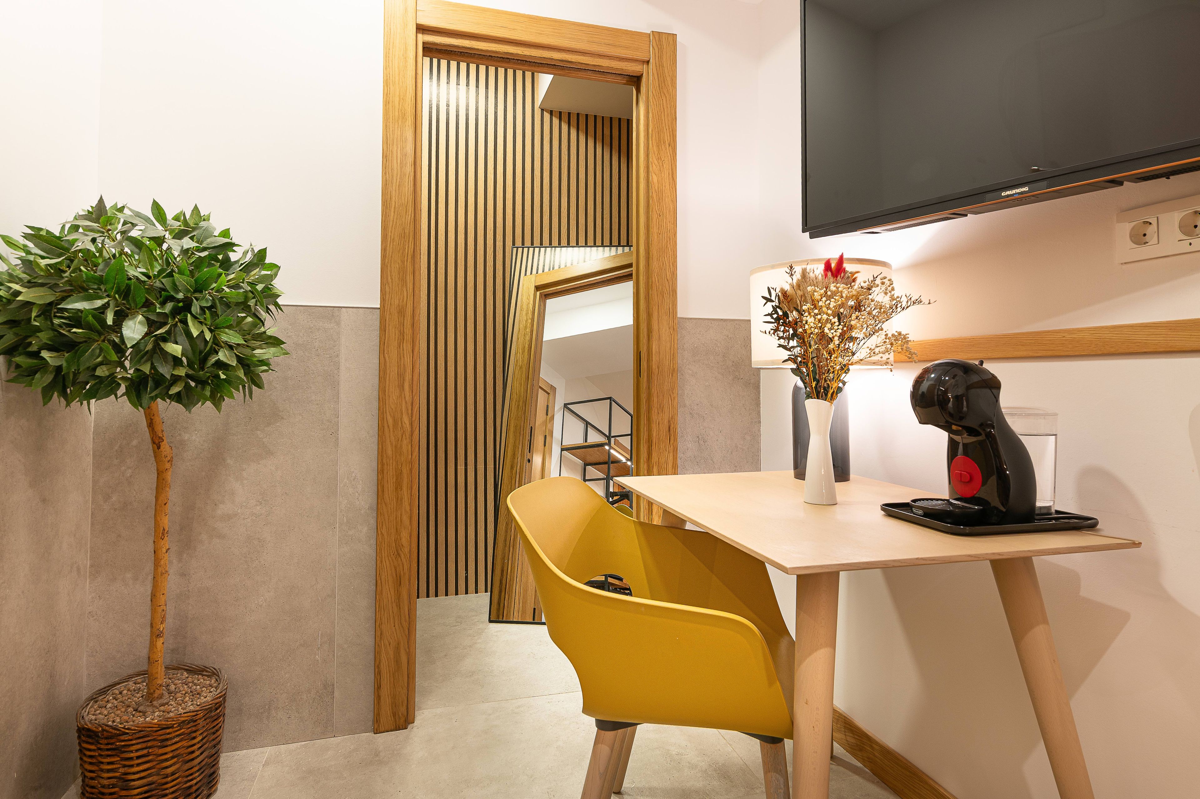 Una habitación con un espejo de marco de madera, una silla amarilla, una mesa con cafetera y una planta en maceta.