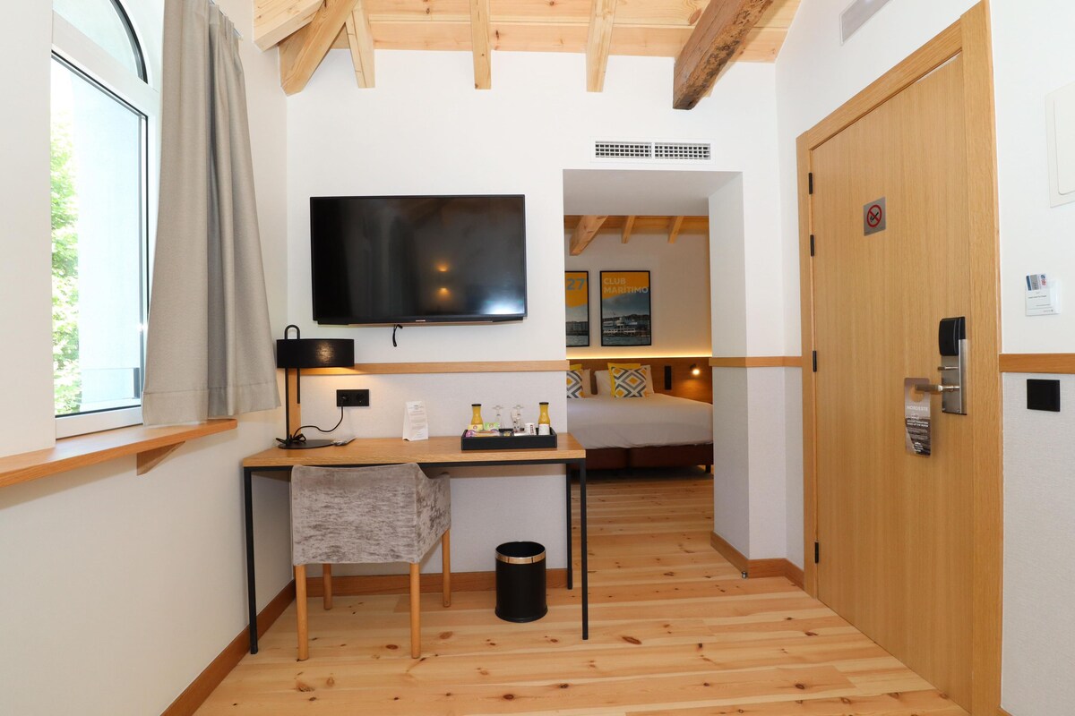 A hotel room with a wooden floor, a desk with a lamp, a television, a bed, and a door with a keycard reader.