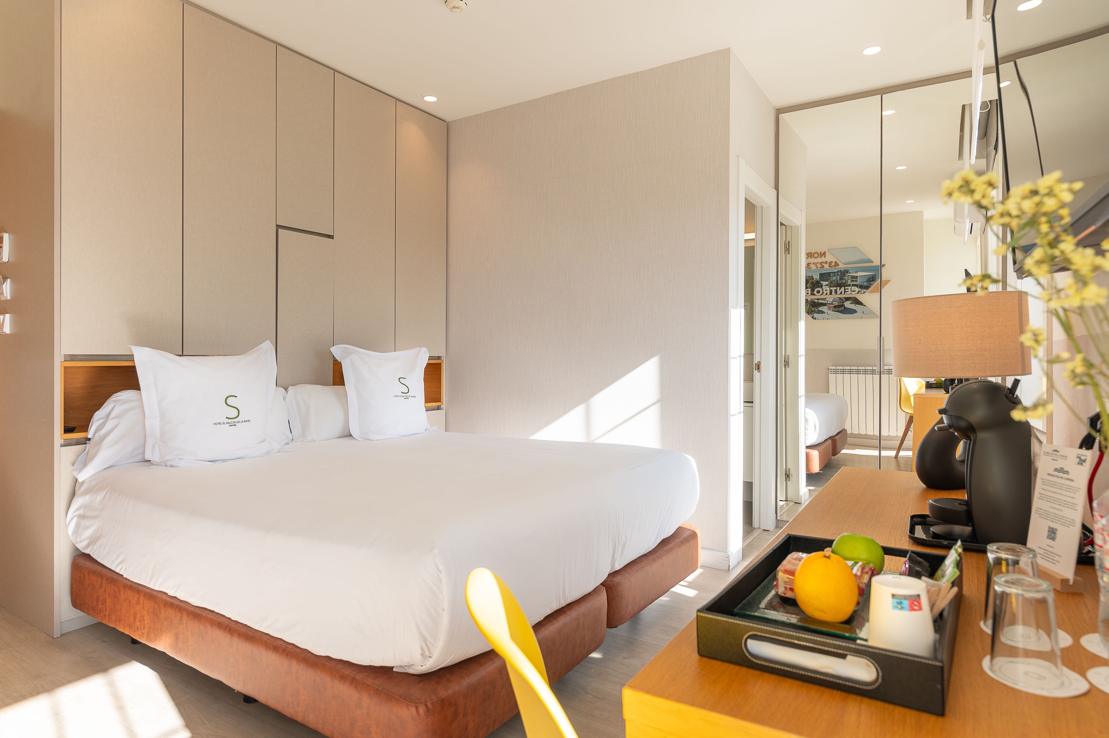 A modern hotel room with a large bed, a wooden headboard, a desk with a coffee maker, and a tray with fruits and a bottle.
