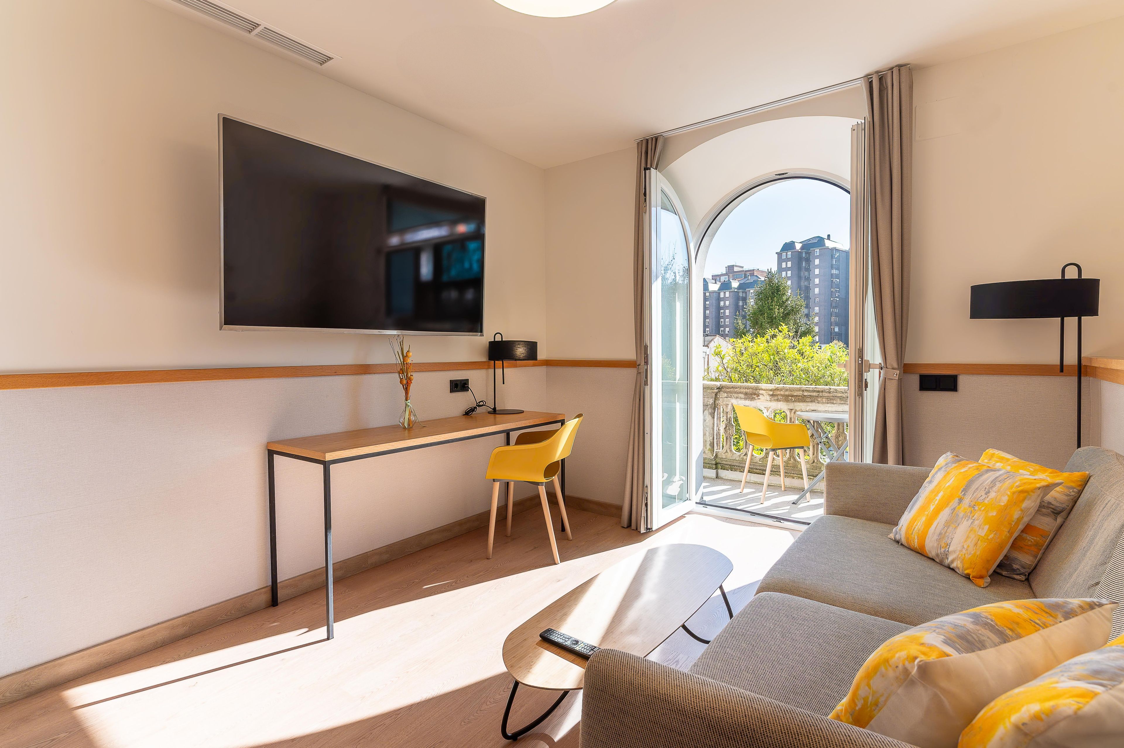 Habitación de hotel con televisión de pantalla plana, un escritorio con silla, sofá con cojines y vistas a edificios desde la ventana.