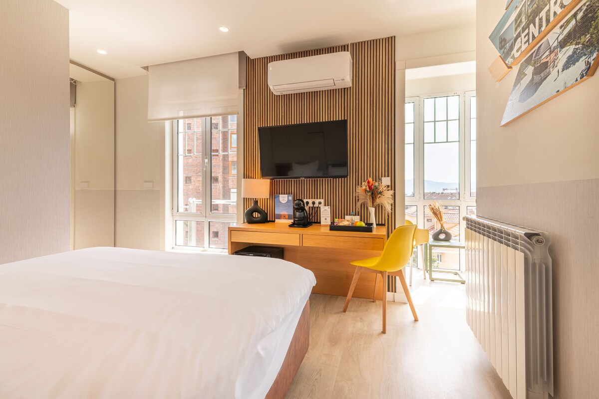 A hotel room with a bed, a desk with a chair, a TV, and a window with a city view.