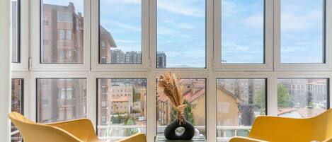 Una habitación con un ventanal grande, dos sillas amarillas, una mesita y un florero con plantas secas.