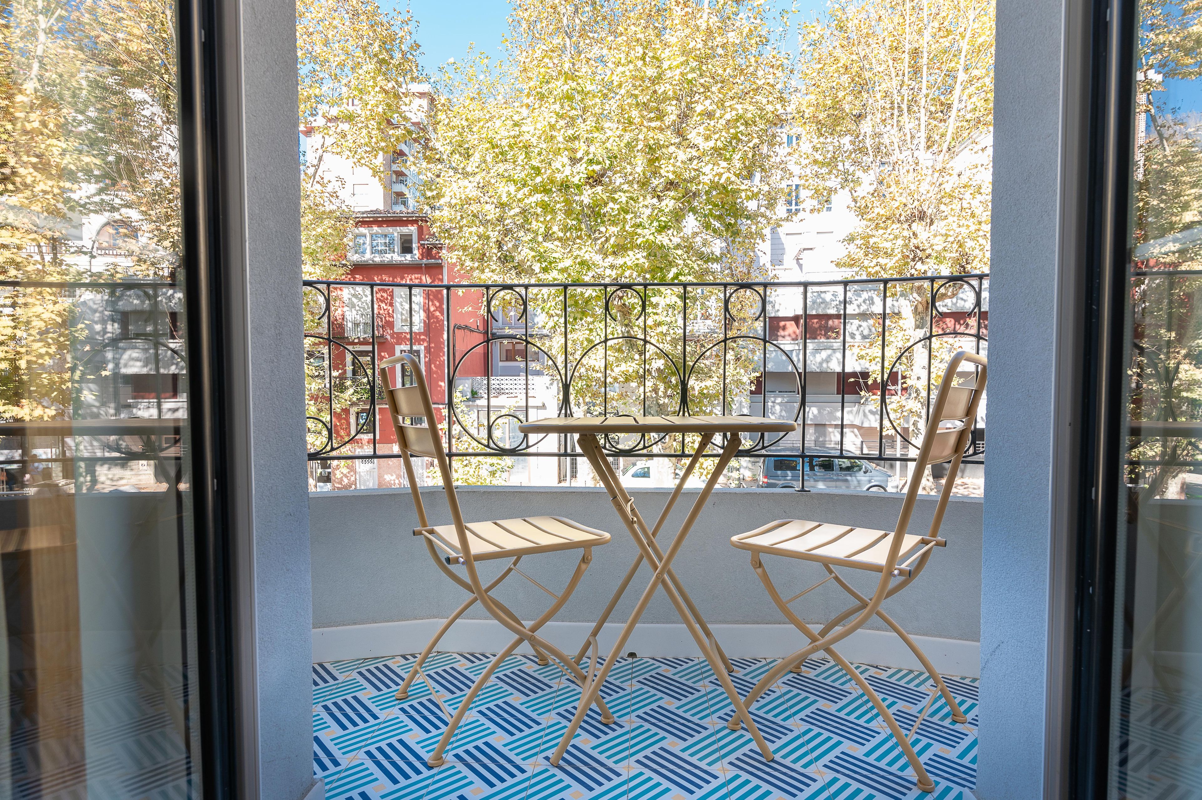 Ein Balkon mit zwei Klappstühlen und einem Tisch, einem gemusterten Boden und Blick auf Gebäude und Bäume.