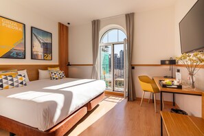 Habitación de hotel con una cama grande, un escritorio con silla, televisor y balcón con vistas a la ciudad.