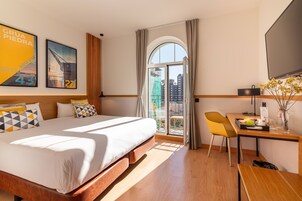Habitación de hotel con una cama grande, un escritorio con silla, televisor y balcón con vistas a la ciudad.