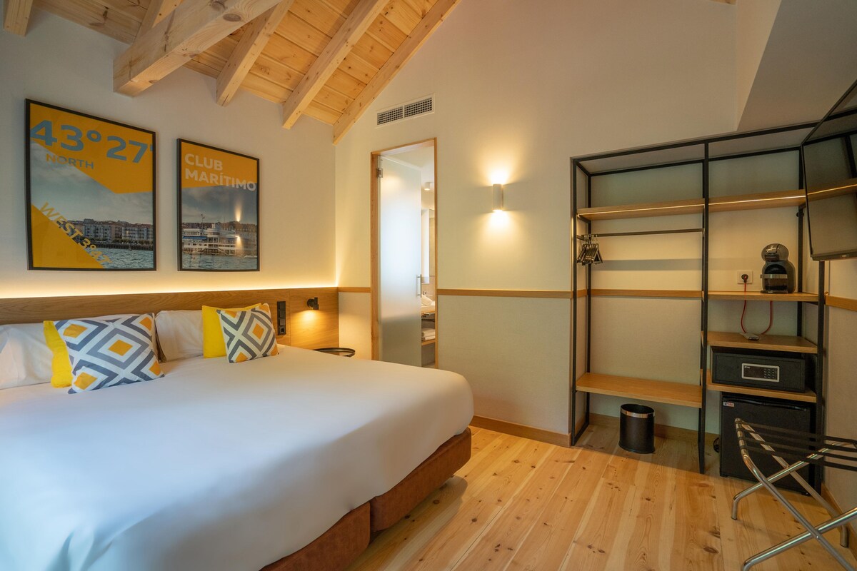 A hotel room with a bed, a desk with shelves, a chair, and a wall-mounted light.