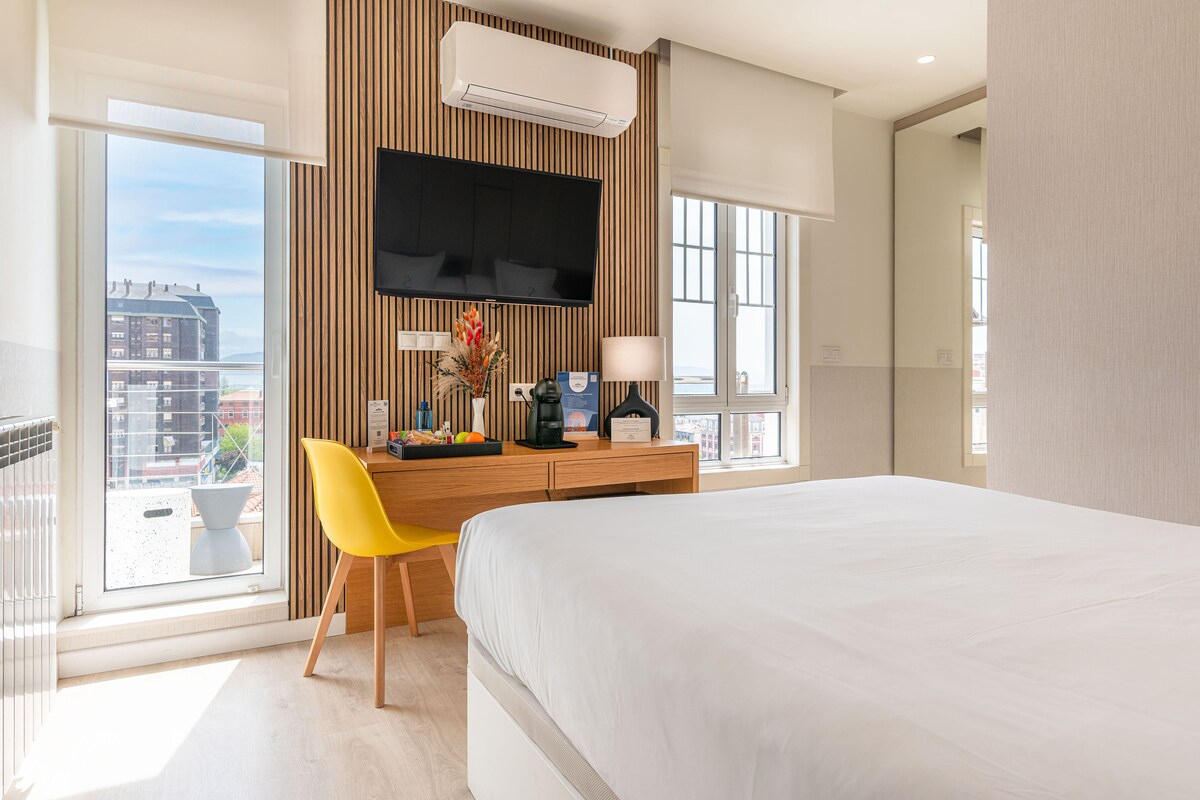 A hotel room with a bed, a desk with a chair, a flat-screen TV, and a view of buildings through large windows.