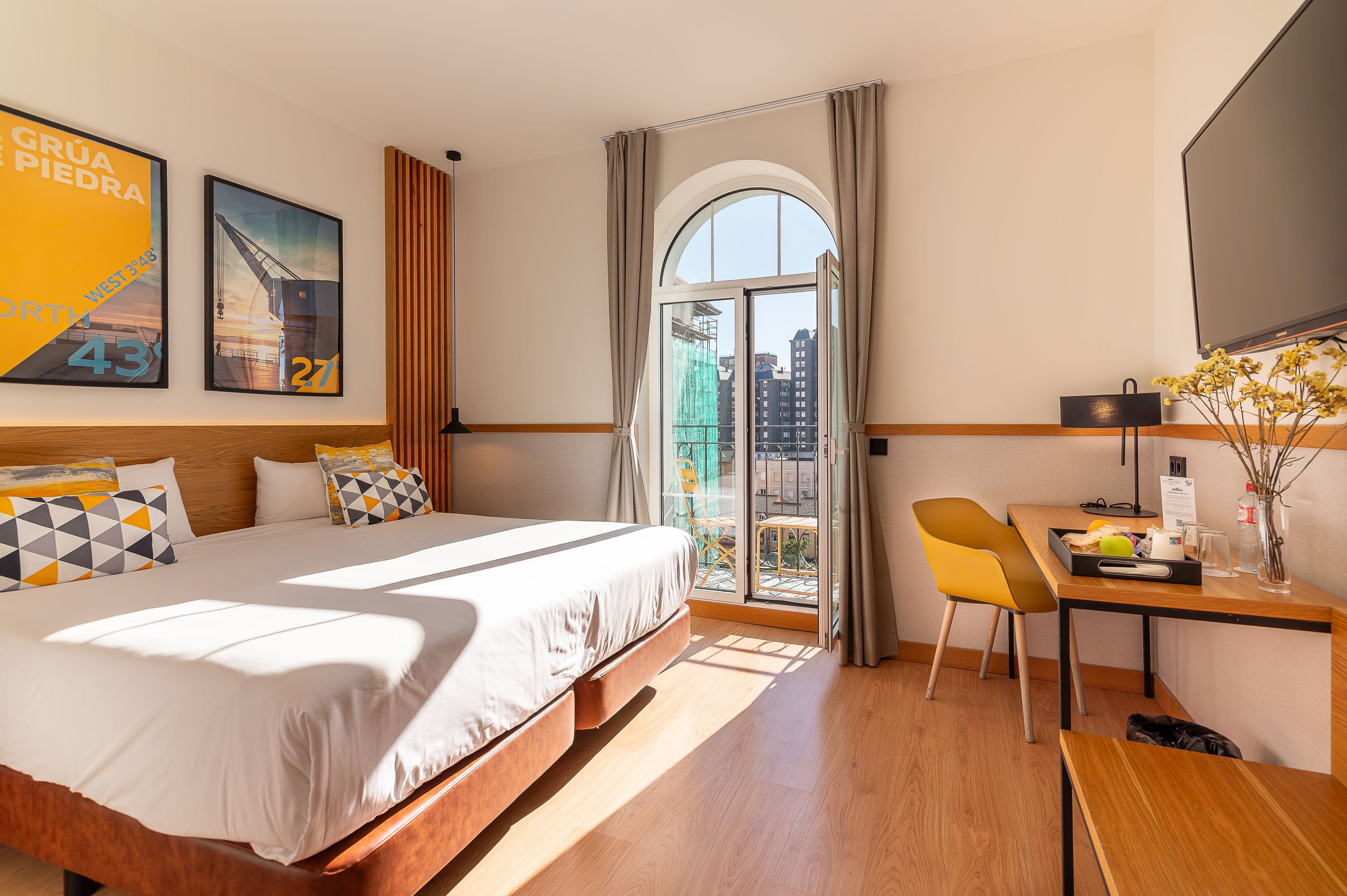 Habitación de hotel con cama, escritorio, silla, ventana con cortinas y vistas a edificios.