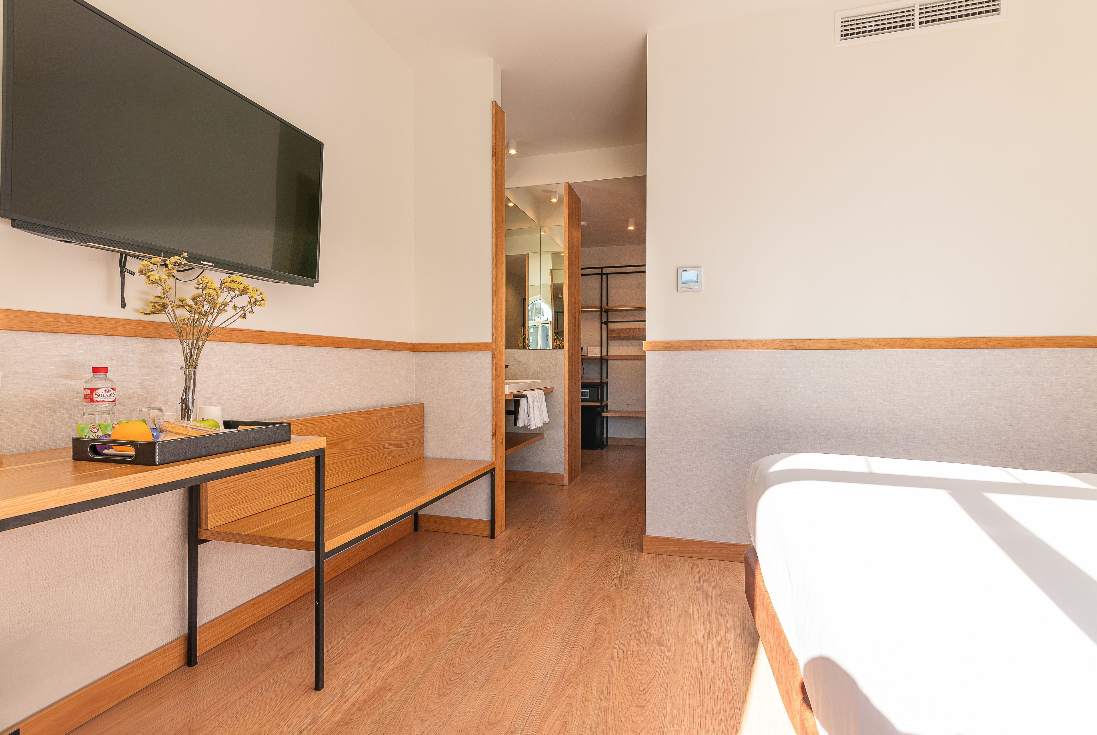 Une chambre d’hôtel comprenant un lit, un bureau avec une chaise, un téléviseur fixé au mur et une petite table sur laquelle est posé un vase de fleurs.