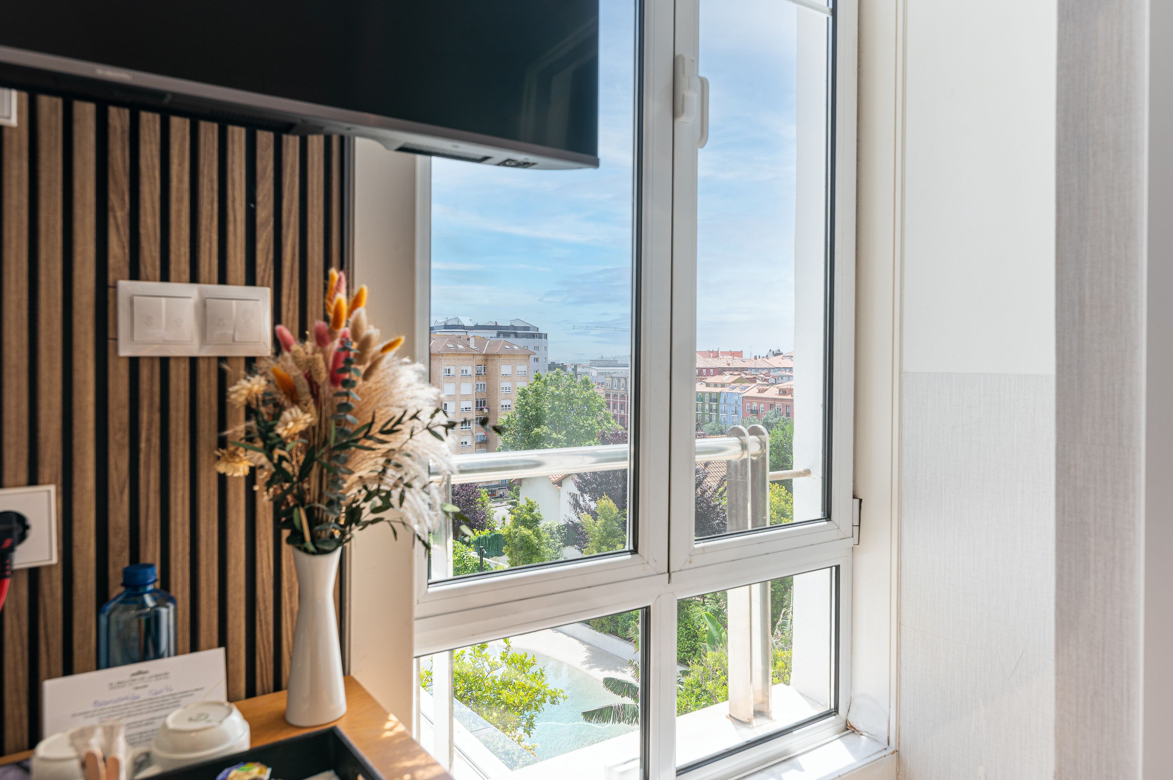 Ein Hotelzimmer mit großem Fenster, einem Flachbildfernseher, einer Vase mit Trockenblumen und einer Wandpaneele aus Holz.