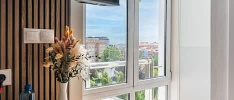 A hotel room with a large window, a flat-screen TV, a vase with dried flowers, and a wooden panel wall.