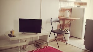 A compact living space with a flat-screen TV on a white desk, a red rug, a chair, and a kitchen area with a refrigerator and a kettle. - Studio Rakella (Opatija)