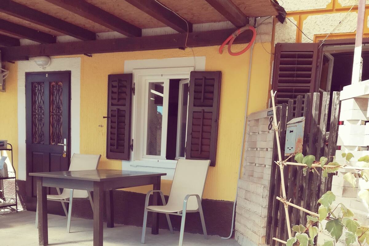 A covered outdoor seating area with a table and chairs, a potted plant, and a door with a decorative design.