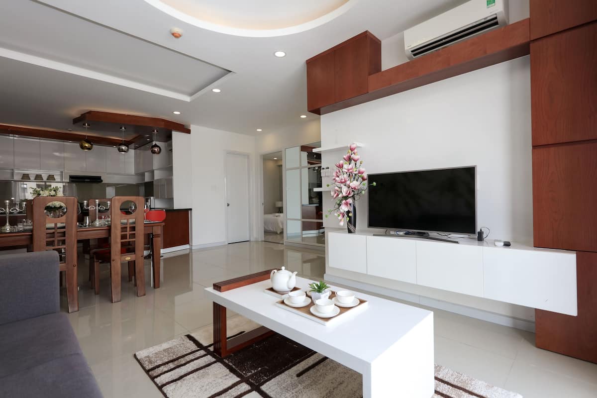A modern living room with a flat-screen TV, a dining area with a wooden table and chairs, and a kitchen in the background.
