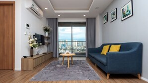 Un salon moderne avec un canapé bleu, un tapis gris, un plancher en bois, un téléviseur posé sur un meuble en bois et une vue sur la ville à travers de grandes fenêtres.