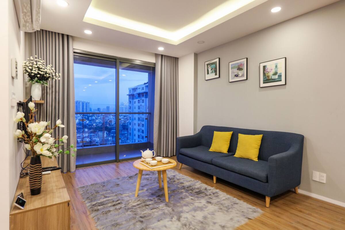 A modern living room with a grey sofa, a small wooden table with a teapot, and a large window with a city view.