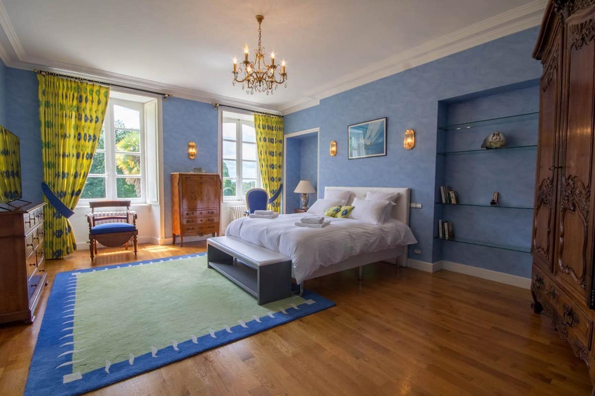 A bedroom with a large bed, a wooden dresser, a chair, a desk, and a bookshelf.