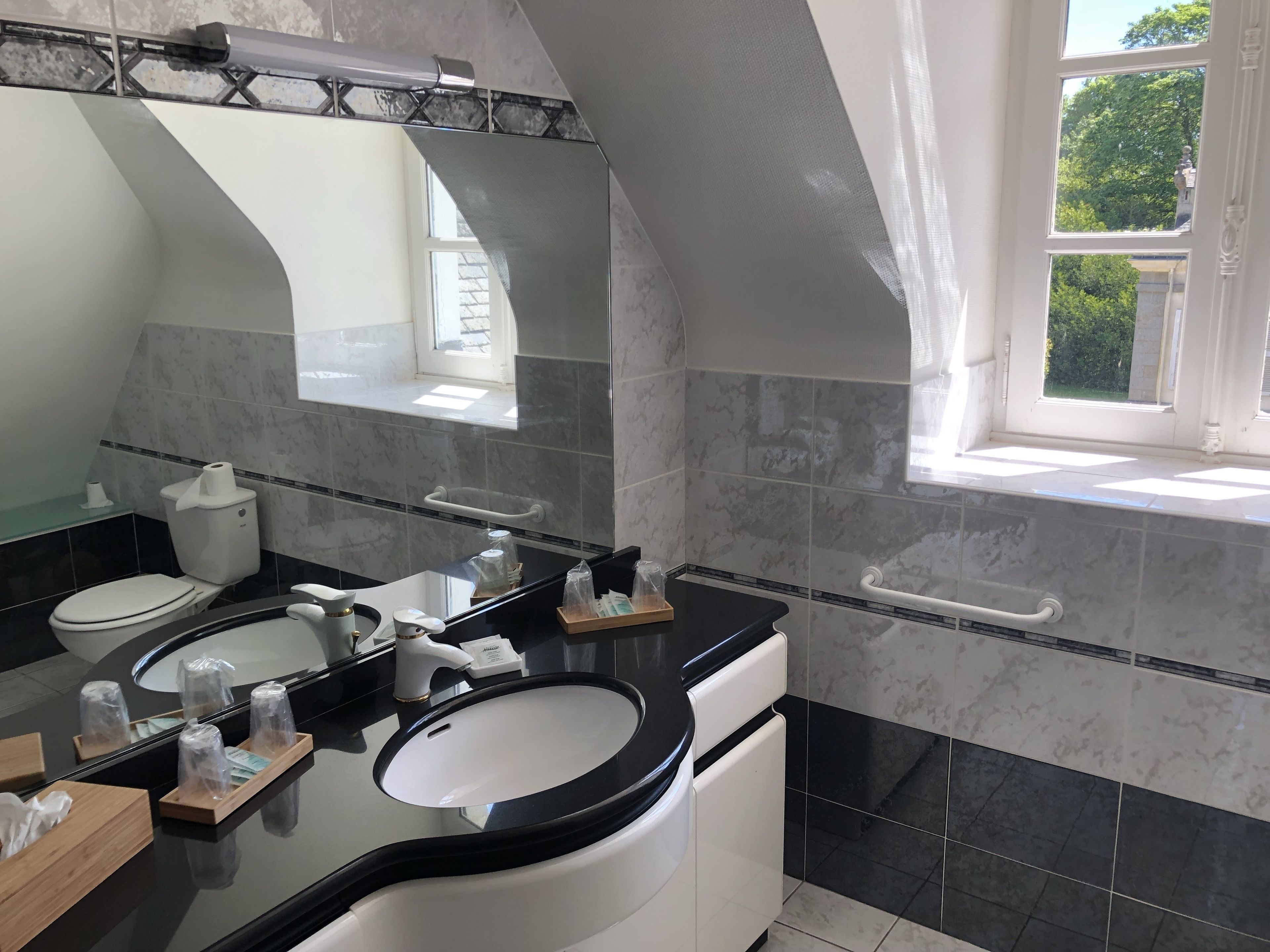 A modern bathroom with two sinks, a toilet, and a bathtub. There is a window providing natural light and a view of greenery outside.