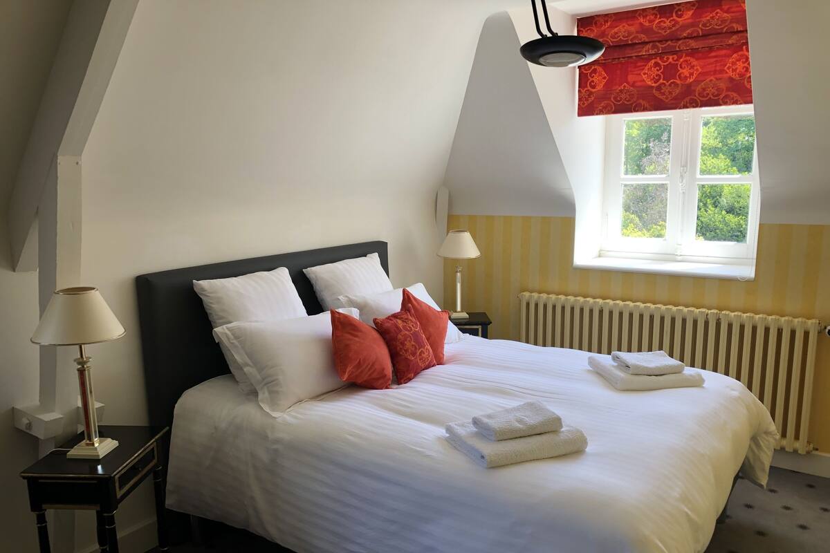 A neatly made bed with white and red pillows, a bedside lamp, and a window with a view of greenery.