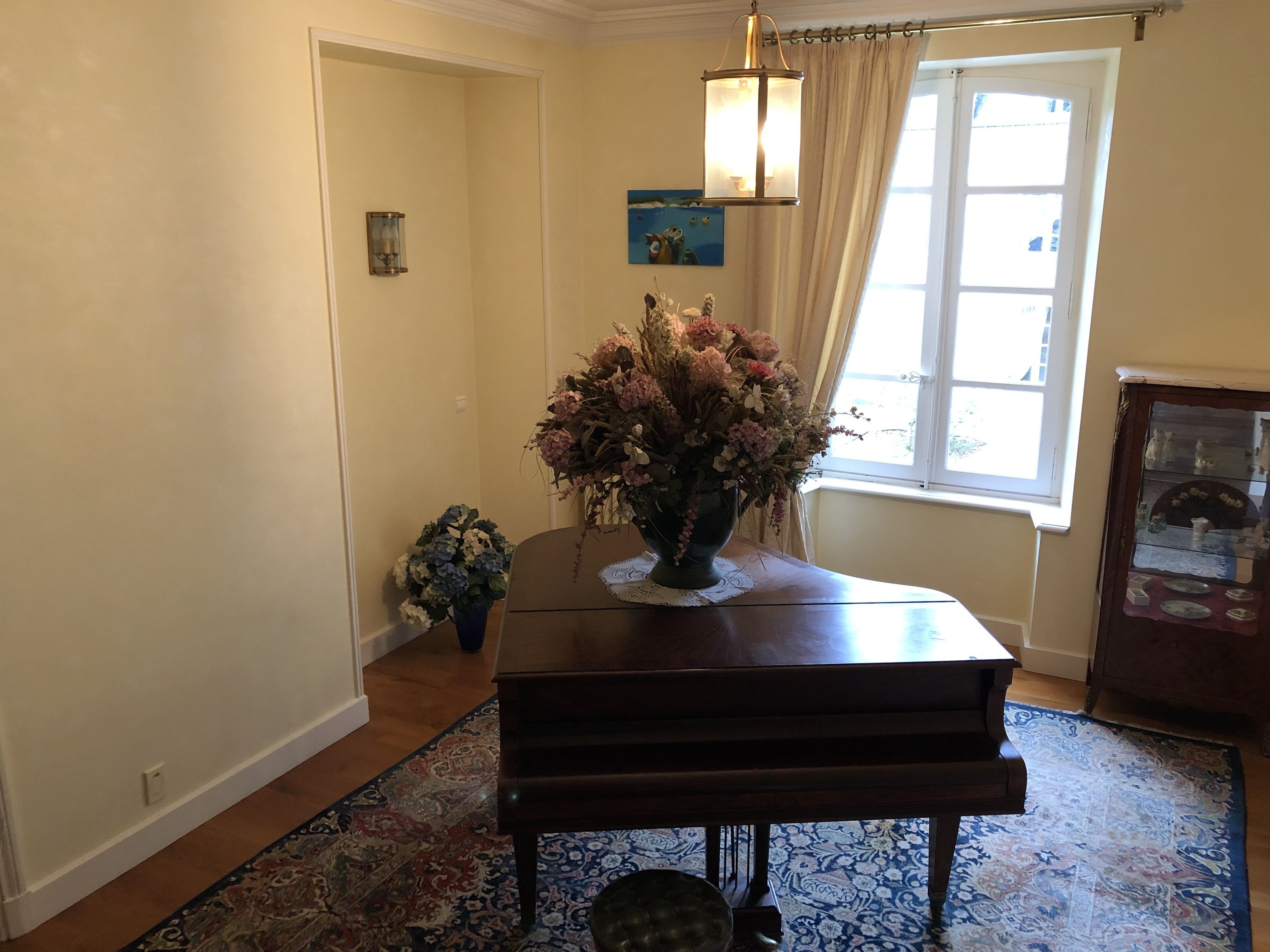 A piano in a room with a window, a vase of flowers, and a patterned rug.