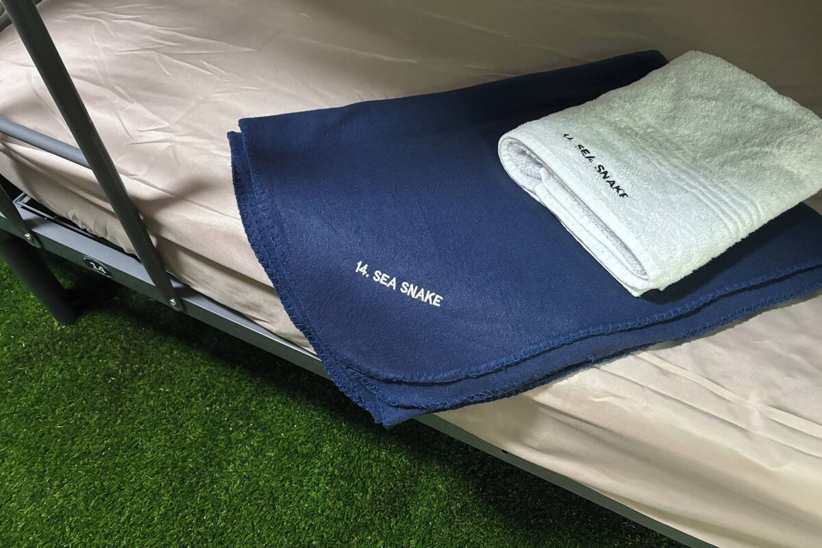 Een bed met een blauwe deken en een handdoek met de tekst 'SEA SNAKE', een metalen frame en een vloer van groen kunstgras.
