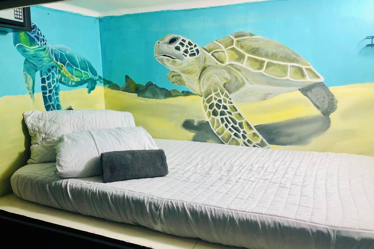 Een bed met een witte matras en kussens, een muurschildering van een zeeschildpad en een bedieningspaneel erboven.