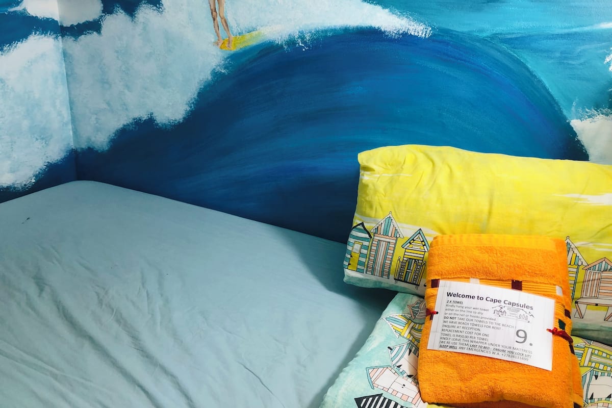 Een slaapkamer met een muurschildering van een surfer op een golf, een bed met een grijs matras en twee kussens met strandmotieven.