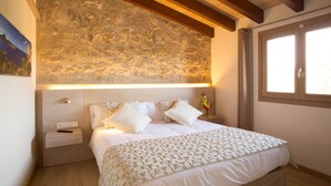 A bedroom with a stone wall, wooden beams, a bed with white bedding, a nightstand, and a window.