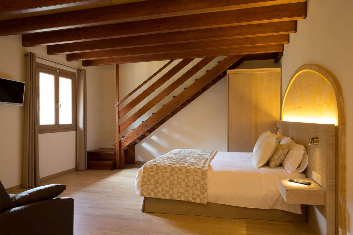 A bedroom with a wooden staircase, a bed with a patterned bedspread, a bedside table with a lamp, and a TV mounted on the wall.