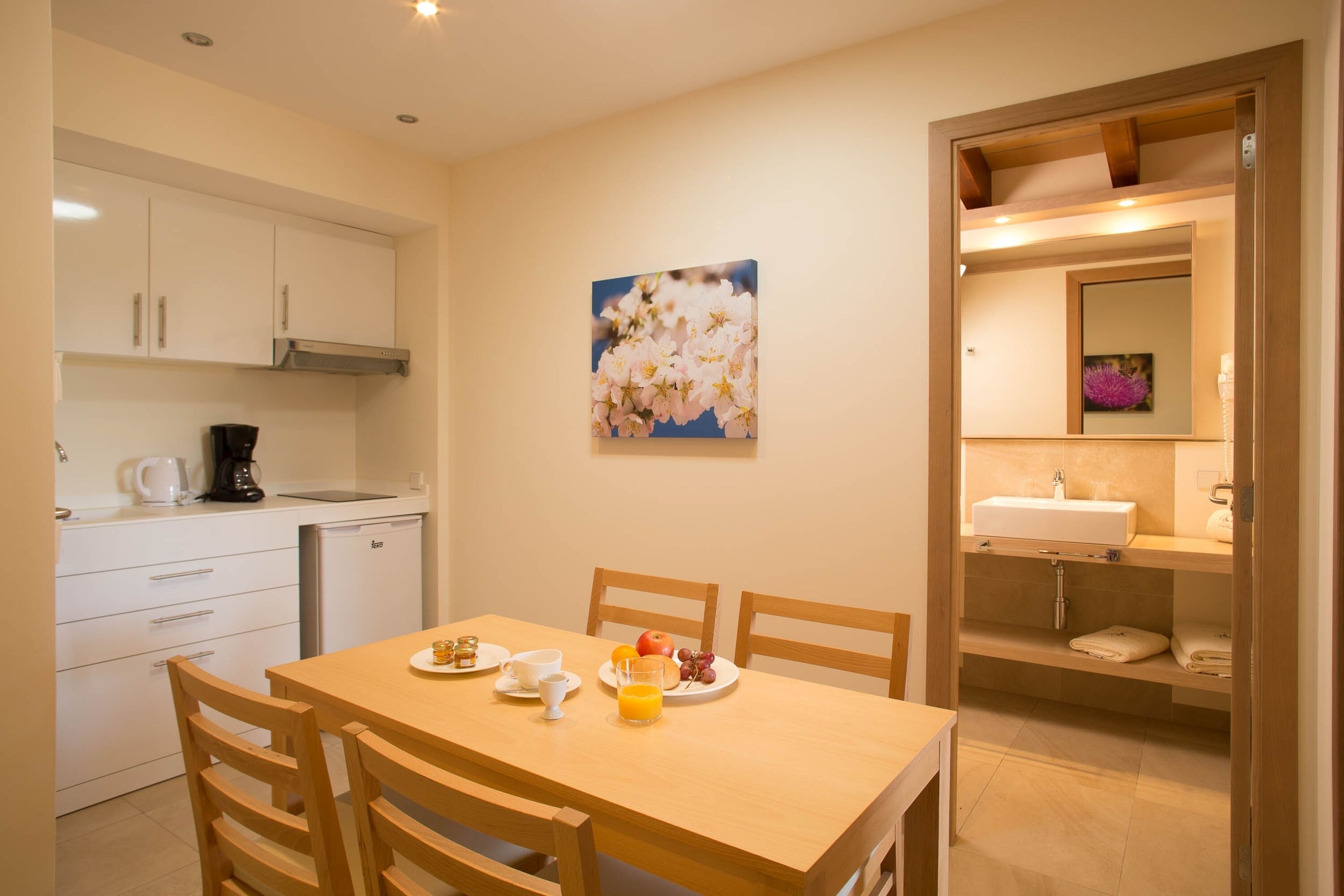 Cocina compacta con armarios blancos, cafetera y mesa de comedor dispuesta con platos, tazas y frutero.