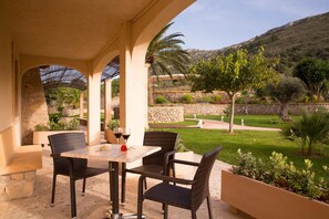 An outdoor patio with wicker furniture and a table, surrounded by stone walls and greenery, overlooking a landscaped garden and hills. - Agroturismo S'Arboçar  (Sant Llorenc des Cardassar)