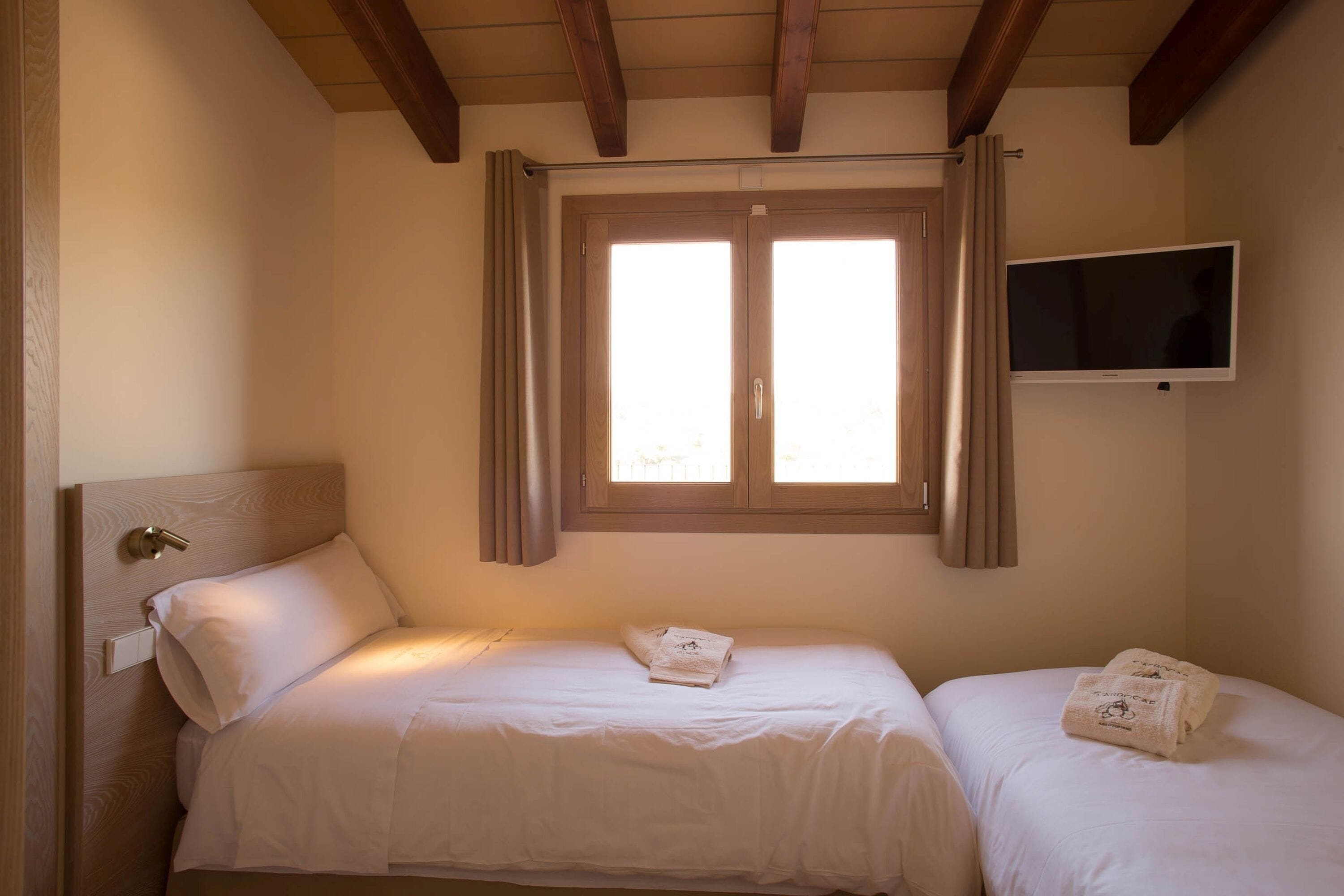A bedroom with two beds, a TV mounted on the wall, and a window with curtains.